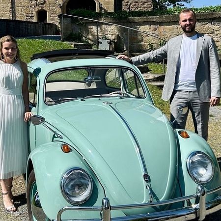 Ein Mann und eine Frau stehen neben einem klassischen VW Käfer und lächeln in die Kamera.