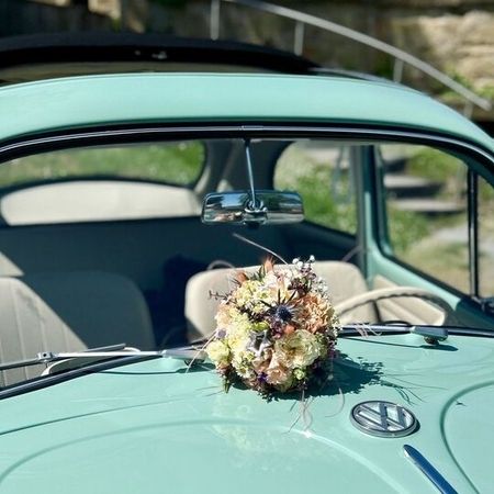 Ein türkiser VW Käfer mit einem Brautstraß auf der Motorhaube, der nostalgische Eleganz und natürliche Schönheit vereint.