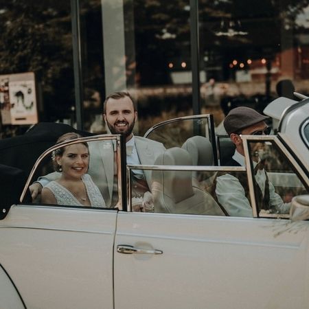 Der VW Käfer Cabrio 1303 steht auf der Straße mit Fahrer und einem Brautpaar auf der Rückbank.