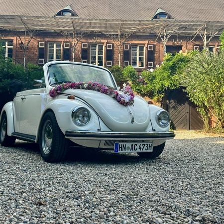Der VW Käfer Cabrio 1303 mit Blumenschmuck auf der Motorhaube steht auf einer Schotterstraße vor einem Haus.