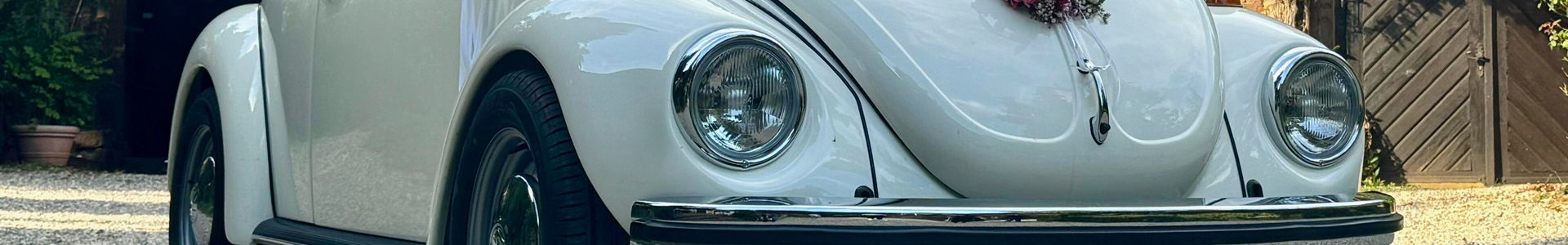 Der VW Käfer Cabrio 1303 mit Blumenschmuck auf der Motorhaube steht auf einer Schotterstraße vor einem Haus.